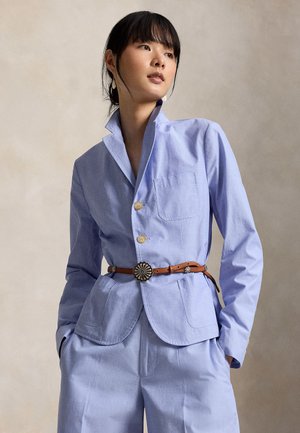 Kobieta ubrana w jasnoniebieski, dopasowany garnitur z brązowym skórzanym paskiem, stojąca z rękami w kieszeniach na neutralnym tle.