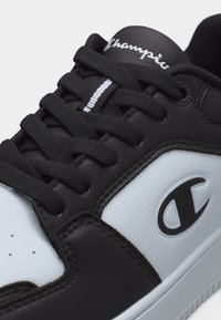 Ασπρόμαυρο δερμάτινο αθλητικό παπούτσι με χοντρές μαύρες κορδόνια, λογότυπο Champion στο πλάι και την επιγραφή «Champion» στη γλώσσα.