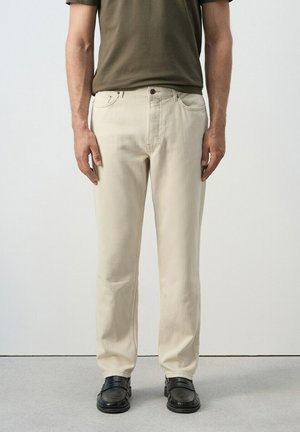 Hombre vistiendo pantalones beige de pierna recta, mocasines negros y una camisa de manga corta verde oliva, de pie contra un fondo liso.