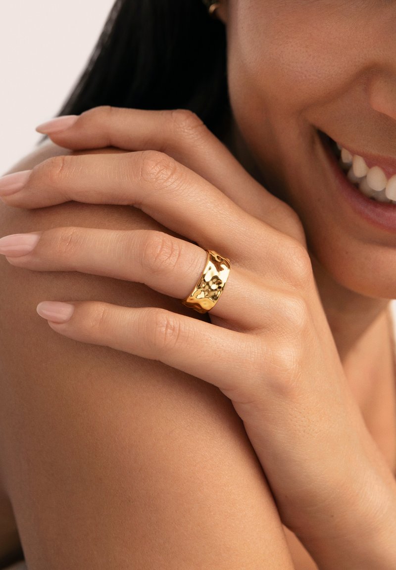 Anillo de oro con superficie texturizada, estilo de banda ancha. Mano reposando sobre la piel, uñas cortas con un ligero esmalte.
