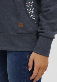 Sweatshirt gris foncé avec une poche à motif floral et un patch en cuir. Il présente des poignets côtelés et une texture douce.