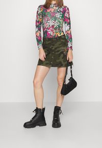 Bloemige bodysuit met lange mouwen in levendige kleuren, gecombineerd met een camouflage denimrok met knopen aan de voorkant en geborduurde zakken. Zwarte combat boots en een kleine zwarte tas maken de look compleet.