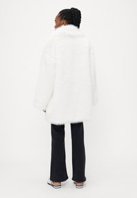 Biely kožúšok z umelej kože s vysokým golierom, dlhými rukávmi a voľným strihom, skombinovaný s čiernymi rozšírenými nohavicami a vzorovanými topánkami.