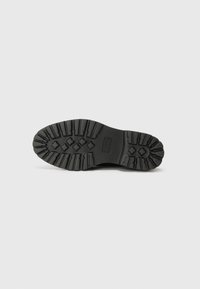 Semelle en caoutchouc noir avec motif de crampons profond, présentant une surface texturée et des accents en forme de diamant pour une meilleure adhérence et durabilité.