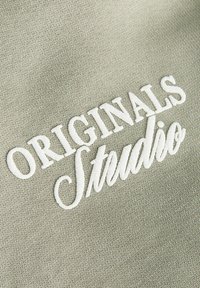 Λευκό κεντητό κείμενο "ORIGINALS Studio" σε φόντο από ελαφρώς γκρι υφασμένο ύφασμα.