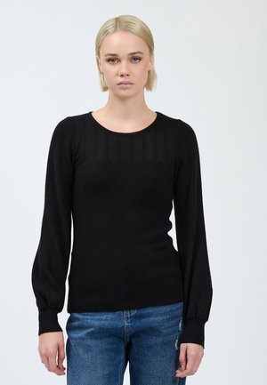 Vrouw met kort blond haar, gekleed in een zwarte gebreide trui met lange mouwen en blauwe jeans, staande tegen een effen lichte achtergrond.