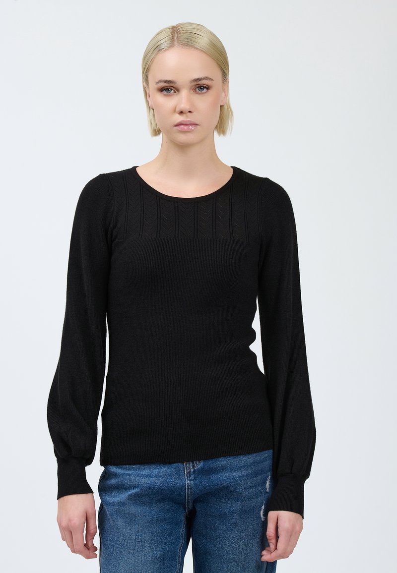 Donna con i capelli biondi corti, che indossa un maglione nero a maniche lunghe e jeans blu, in piedi di fronte a uno sfondo chiaro e semplice.