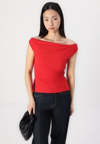 Red off-the-shoulder top made of smooth, stretchy fabric with gathered side detail. Paired with dark blue wide-leg jeans and a black handbag.