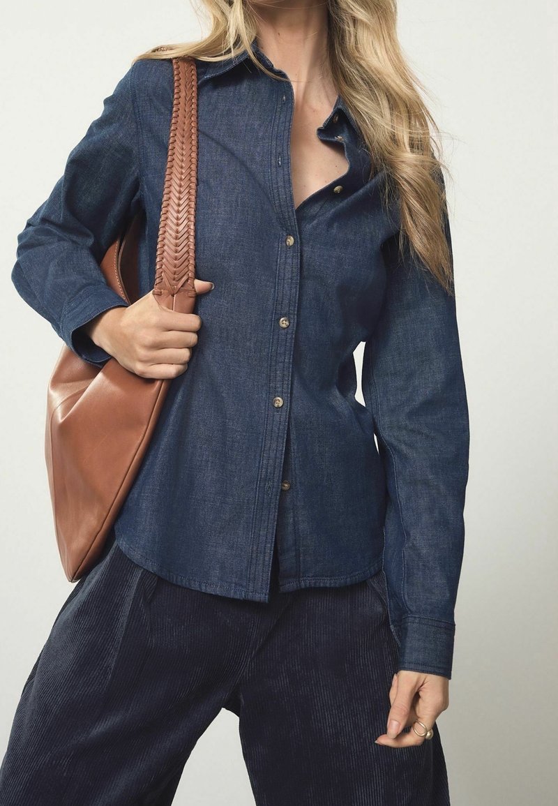 Femme portant une chemise en jean foncé boutonnée et un pantalon sombre, portant un grand sac à bandoulière en cuir marron avec une sangle tressée.