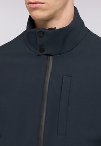 Giacca blu scuro con colletto alto, dotata di chiusura a bottoni a scatto, zip frontale e dettaglio della tasca rettangolare. Tessuto liscio.