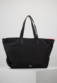 Grand sac fourre-tout noir avec double poignée en cuir, doublure intérieure rouge et petit logo métallique Karl Lagerfeld à l'avant, posé sur une surface blanche.