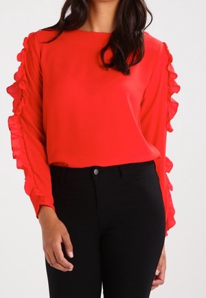 Vrouw draagt een felrode blouse met lange mouwen en ruches aan de mouwen en zwarte hoge taille broek tegen een effen achtergrond.