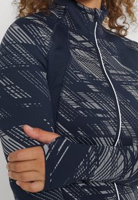 Marinblå mönstrad zip-jacka med grå diagonala linjer, med hög krage och åtsittande ärmar med tumhål. Slät, stretchig tyg.