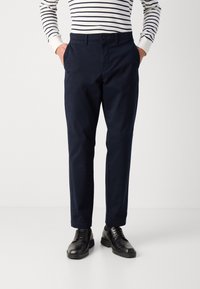 Mörkblå bomullsbyxor med rak skärning, två frontfickor och knäppning fram, tillsammans med svarta läderskor.