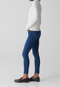 Jeans skinny in denim blu scuro, con vita media e dettagli di cucitura minimi. Abbinati a un top bianco a maniche lunghe e mocassini marroni.