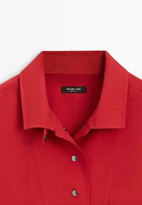 Chemise rouge à col avec boutons-pression noirs et étiquette "Massimo Dutti" visible à l'intérieur du col.