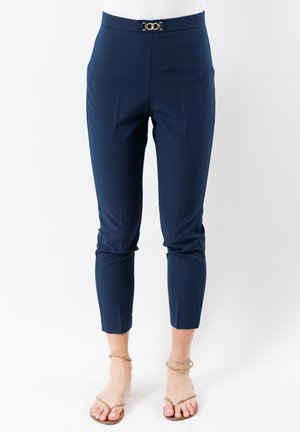 Pantaloni alla caviglia blu navy su misura in tessuto liscio, con una fibbia frontale in tonalità oro e pieghe frontali ben definite.
