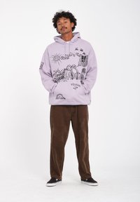 Un sweat à capuche lilas avec un design graphique représentant la nature et des figures, associé à un pantalon en velours côtelé marron. Tissu texturé, coupe décontractée et poche kangourou.