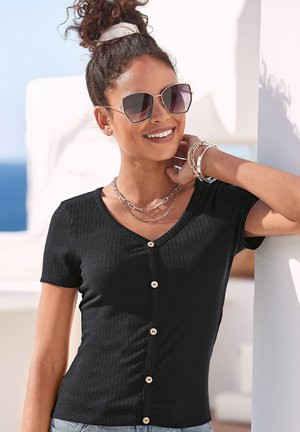 Lächelnde Frau mit Sonnenbrille, schwarzem, geripptem Hemd mit Knöpfen, geschichteten Halsketten, Armbändern, die Haare mit einem Schal zusammengebunden, lehnt sich an eine weiße Wand im Freien.