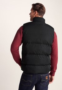 Black puffer vest featuring a high collar and a quilted design, worn over a maroon long-sleeve shirt, paired with dark denim jeans.