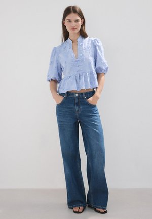 Mujer con blusa texturizada azul claro con mangas abullonadas y vaqueros azules de pierna ancha, de pie con las manos en los bolsillos.