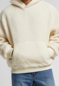 Hoodie in pile color crema, morbido e peloso, con tasca a marsupio frontale, polsini a coste e cappuccio con cordino. Design casual e caldo.