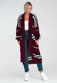 Sweter kardigan w kolorze bordowym, z czarnymi, białymi i beżowymi paskami. Otwarty krój z długimi rękawami, zestawiony z spodniami z wysokim stanem i białymi sneakersami.