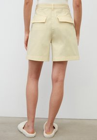 Kvinna som bär krämfärgade shorts med hög midja och fickor med klaff på baksidan, kombinerade med vita fluffiga slip-on skor, stående på grått golv.