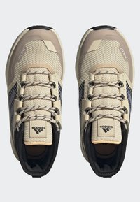 Zapatillas deportivas beige con tejido de malla, acentos negros y suelas estriadas. Los detalles reflectantes y los cordones texturizados completan el diseño.