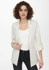 Blazer bianco su misura con maniche a tre quarti, un solo bottone e vestibilità slim, indossato sopra un top bianco a righe e jeans scuri.