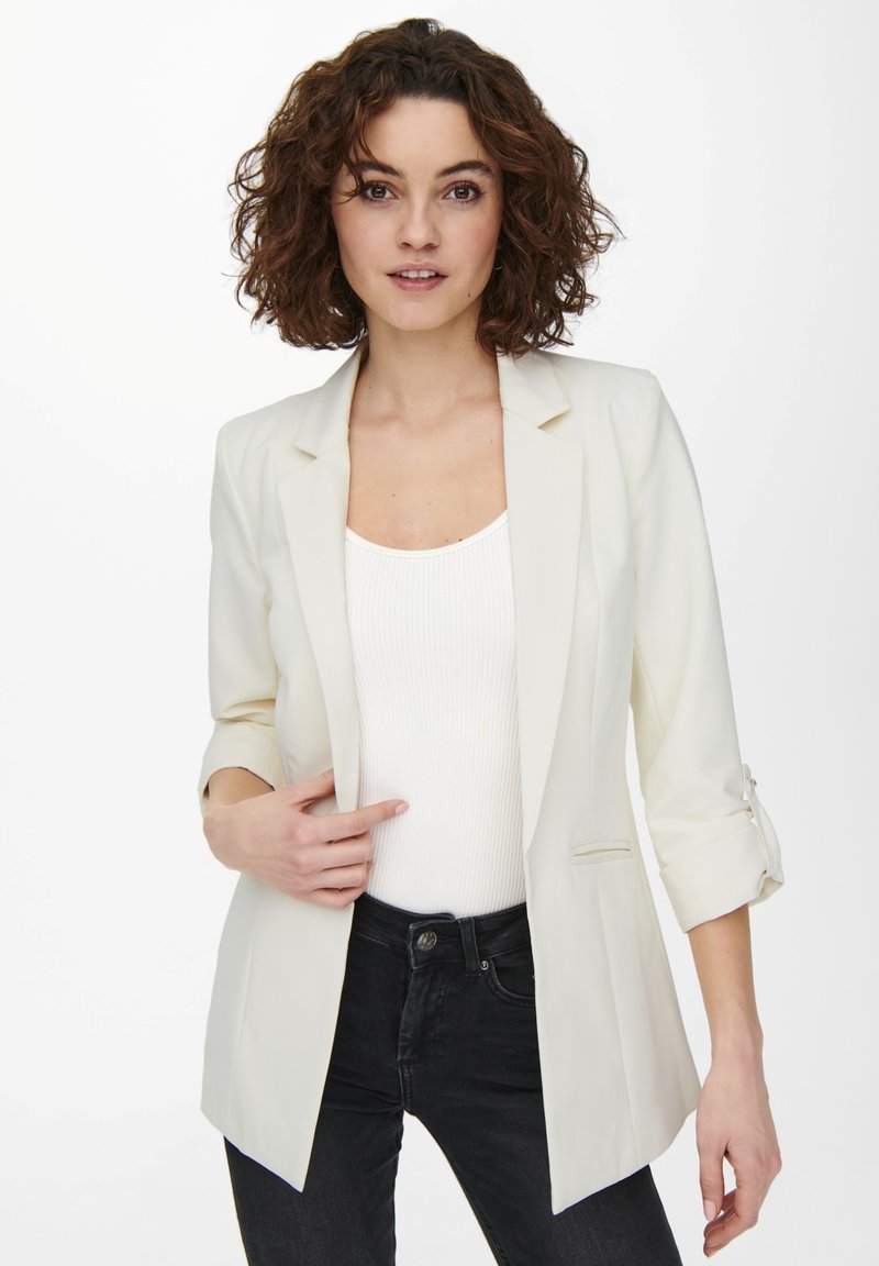 Blazer bianco su misura con maniche a tre quarti, un solo bottone e vestibilità slim, indossato sopra un top bianco a righe e jeans scuri.