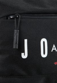 Sac en tissu noir avec le texte blanc "J O A" et un accent rouge. Comprend une fermeture éclair avec un tirant texturé noir, affichant un logo Jordan.