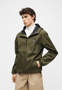 Νεαρός με σγουρά μαλλιά φορώντας μια χακί ζακέτα με φερμουάρ πάνω από έναν λευκό ζιβάγκο και μπεζ παντελόνι, με το ένα χέρι στην τσέπη.