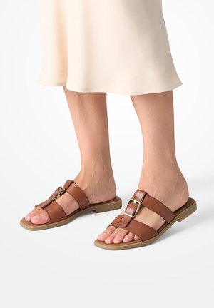 Pies con sandalias de cuero marrón tipo slide con hebillas doradas y una falda beige visible desde la mitad de la pantorrilla hacia abajo.