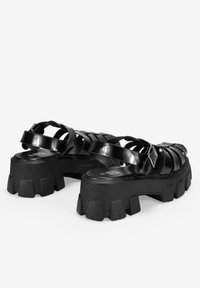 Sandalias de plataforma negras con una suela gruesa, que presentan correas entrelazadas y un detalle de hebilla. El material parece ser brillante y sintético.