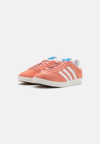 Sneaker in suede rosa con design a tre strisce bianche, dotate di suole in gomma bianca e dettagli blu sulla linguetta.