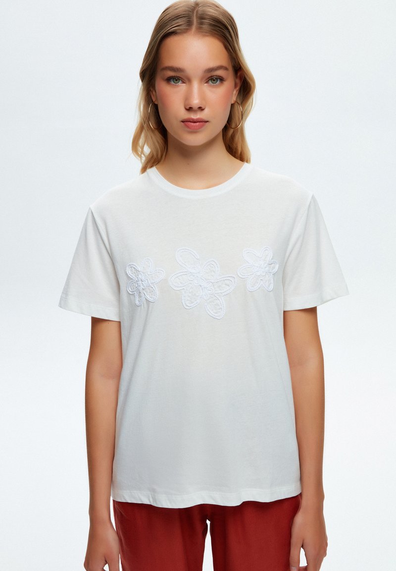 Camiseta de algodón blanca con tres diseños de flores bordados en blanco en el pecho, mangas cortas y escote redondo.