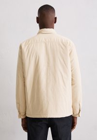 Beżowa bawełniana kurtka o luźnym kroju, z zapiętymi na guziki mankietami i kołnierzem typu wierzchołek, charakteryzująca się gładką fakturą i minimalistycznym designem.