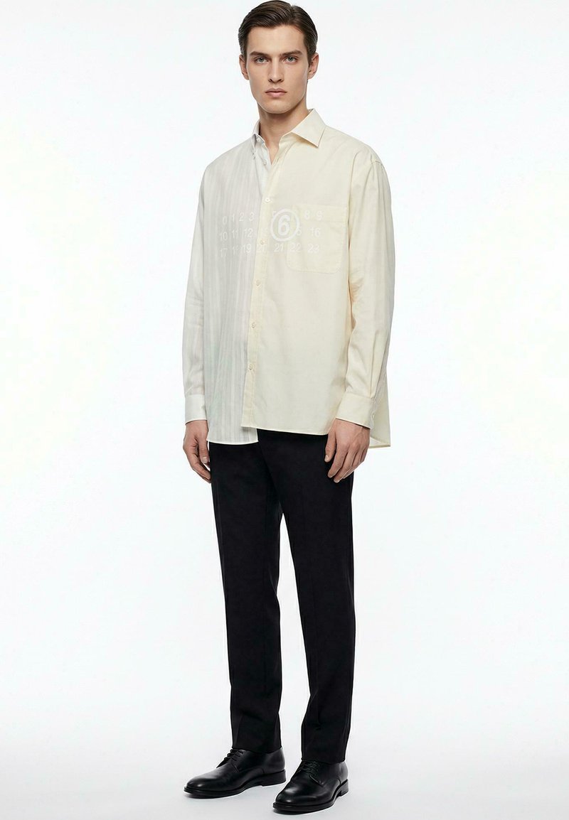 Giovane uomo che indossa una camicia crema a maniche lunghe con bottoni, pantaloni neri e scarpe eleganti nere, in piedi davanti a uno sfondo bianco neutro.