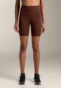 Barna, magas derekú biciklissort nyújtó anyagból, sima felülettel és combközép hosszúsággal, fekete sportcipővel párosítva.