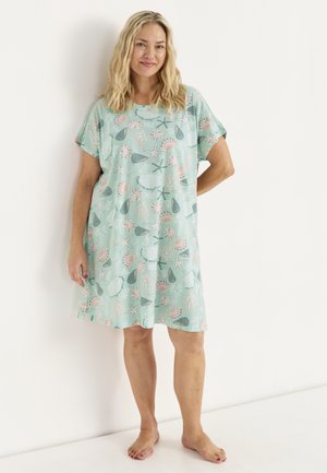 Vrouw die blootsvoets staat en een losse, lichtblauwe jurk draagt met roze en groene schelpen- en zeesterrenpatronen tegen een witte achtergrond.