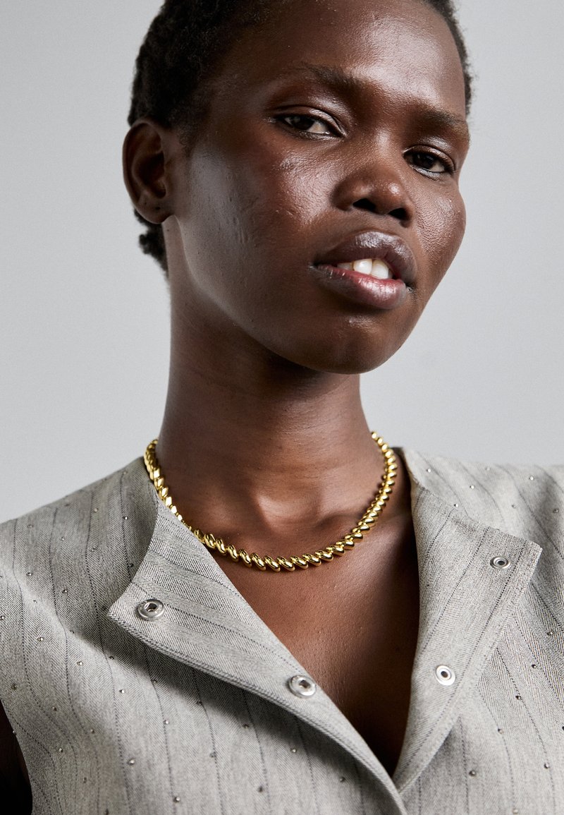 Gilet grigio a spina di pesce con accenti metallici, caratterizzato da una chiusura a doppi bottoni. La collana a catena dorata intrecciata mette in risalto il décolleté.