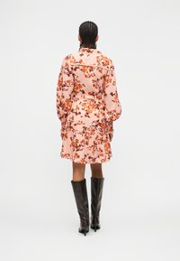 Γυναίκα με πλεξούδες, που φοράει ένα ροζ φόρεμα με λουλούδια και μαύρες μπότες με τακούνι μέχρι το γόνατο, στέκεται με την πλάτη προς το μέρος μας σε λευκό φόντο.