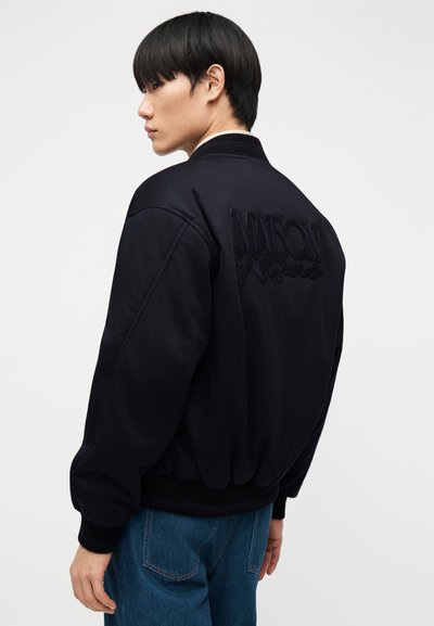 Junger Mann im Profil, der eine schwarze Bomberjacke mit gesticktem "MAISON"-Schriftzug auf dem Rücken und blaue Jeans trägt, vor einfachem Hintergrund.