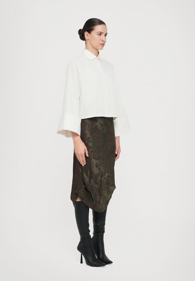 Weißes, kmissiertes Oberteil mit Kragen und Glockenärmeln, kombiniert mit einem knielangen, olivgrünen Satinrock mit floralem Muster. Schwarze Stiefel bis zur Oberschenkelmitte.