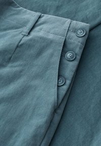 Zbliżenie na turkusowe spodnie w korduroy z trzema widocznymi guzikami zapinającymi pas i szlufkami na pasek.