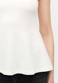 Κοντινό πλάνο στον κορμό ενός ατόμου που φοράει λευκό αμάνικο τοπ με φουσκωτή άκρη και σκούρο παντελόνι, πάνω σε απλό φόντο.