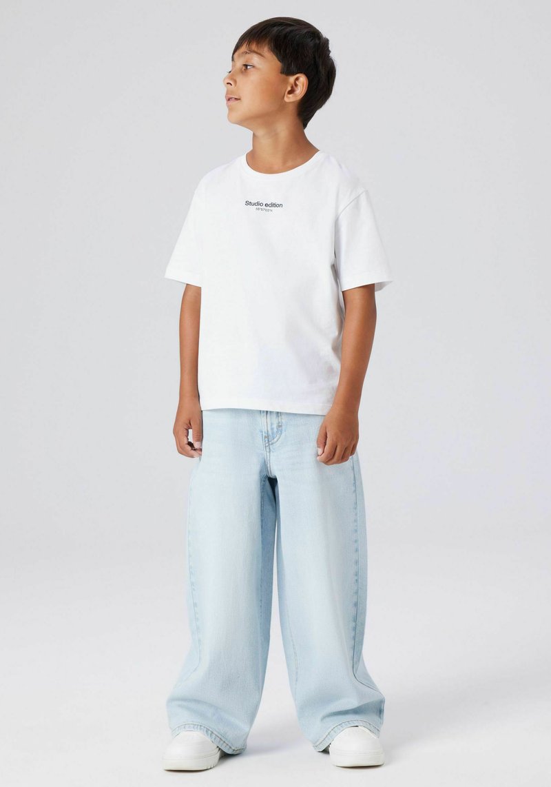 Balta īsroku T-krekls ar uzrakstu "Studio edition", kopā ar gaiši zilām plaša piegriezuma džinsa biksēm un baltām sporta apaviem. Ikdienišķs piegrieziens un relaksēts stils.