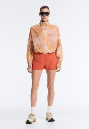 Ženska, ki nosi pretirano veliko breskovo jakno, oranžne kratke hlače, bele nogavice, bež superge in črna sončna očala, stoji na belem ozadju.