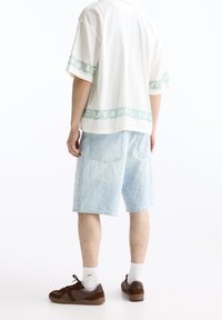 Hombre con camisa blanca de manga corta con bandas de patrón verde claro, pantalones cortos de mezclilla azul claro con estampado, calcetines blancos y zapatillas marrones, de pie.